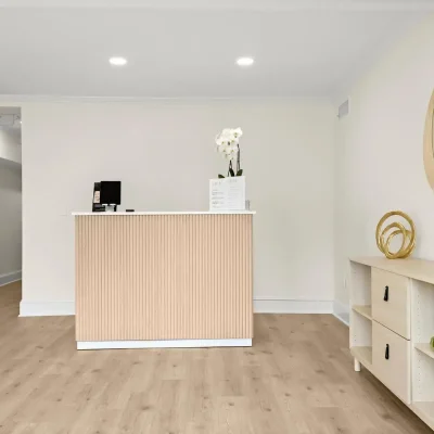 Spacious reception area of a facial spa with a wooden front desk, minimalist shelving with plants and decor, and a spacious, brightly lit room with wooden flooring.